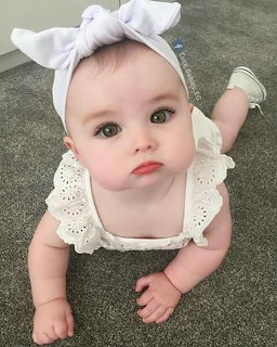 Baby wearing a white bow headband and lace outfit crawling on a gray carpet