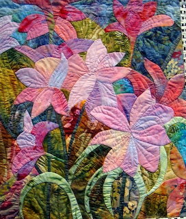 Quilted fabric artwork of pink and purple lilies with green leaves in patchwork textile art