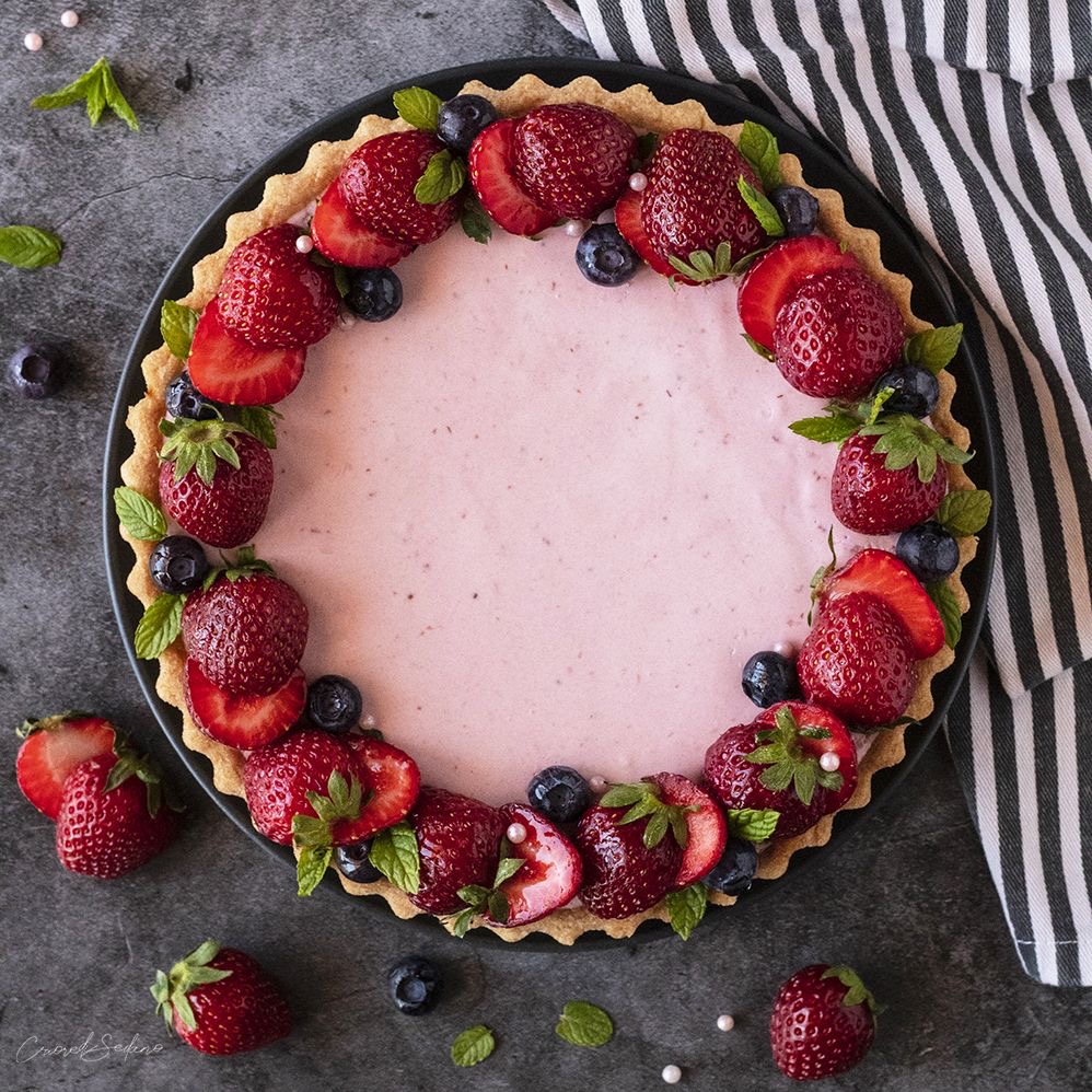 Overhead view of a pink tart topped with strawberries and blueberries on a dark table