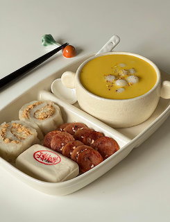Cream tray with yellow pumpkin soup in a pot, rice cakes, stuffed rolls, and Korean snacks with chopsticks