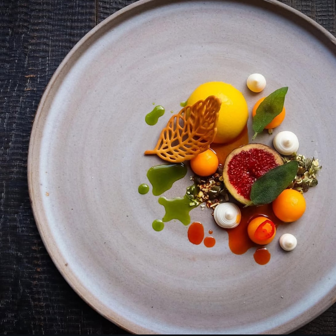 Artistic plated dessert with yellow sphere, fig, kumquats, and white meringues on gray ceramic plate