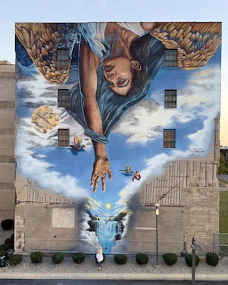 Vertical mural of woman in blue shirt reaching up, painted on brick building with cloudy sky background