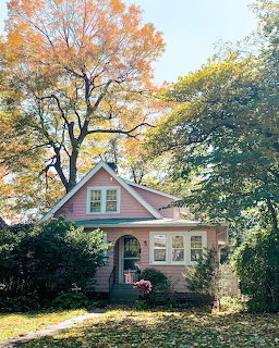 Pink cottage with white windows surrounded by tall trees with autumn foliage
