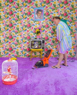 Man in striped robe vacuuming vivid purple carpet in a colorful room with floral wallpaper and vintage TV