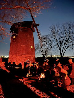 Colonial musicians in tricorn hats playing fifes and drums at dusk beside an illuminated red windmill