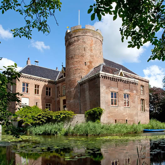 Red brick castle with cylindrical tower beside moat with water lilies, framed by tree branches