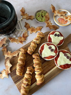 Halloween party food with mummy hot dogs, ghost mini pizzas, bat chips, and guacamole on a cutting board
