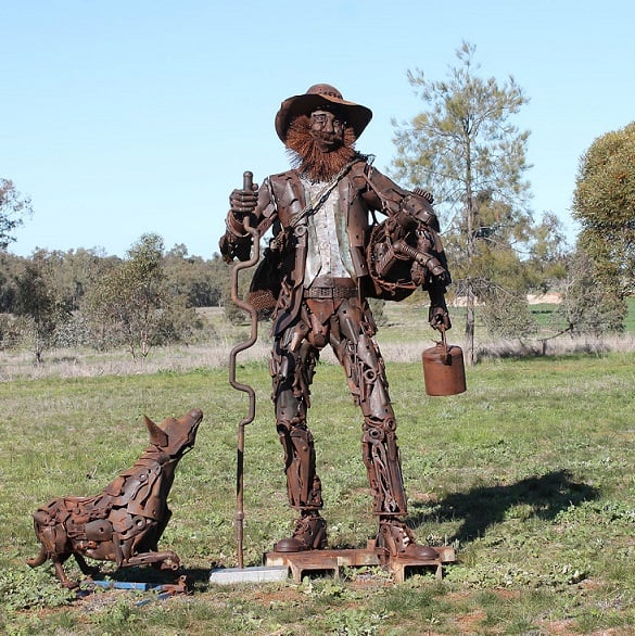Rusty metal swagman sculpture with a seated dog on grass outdoors