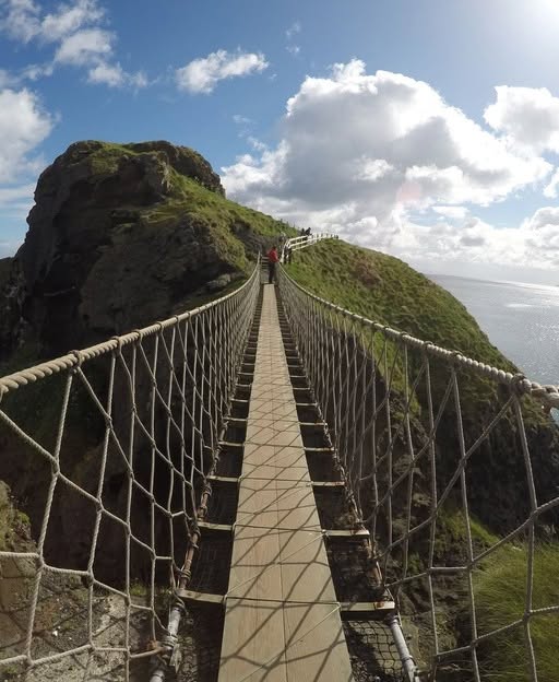 Rope bridge connecting green coastal cliffs over ocean gap with blue sky and clouds