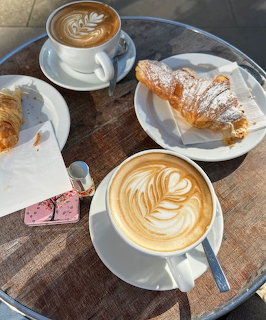 Overhead view of a cafe table with two lattes featuring leaf art, croissants, and a pink wafer cookie