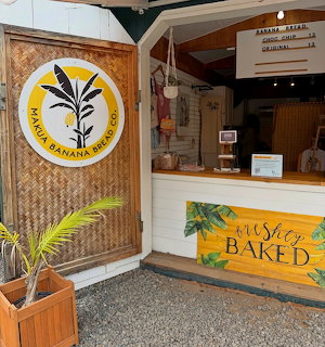 Maui Banana Bread Co. shop stall with round logo sign, wooden counter, and freshly baked sign