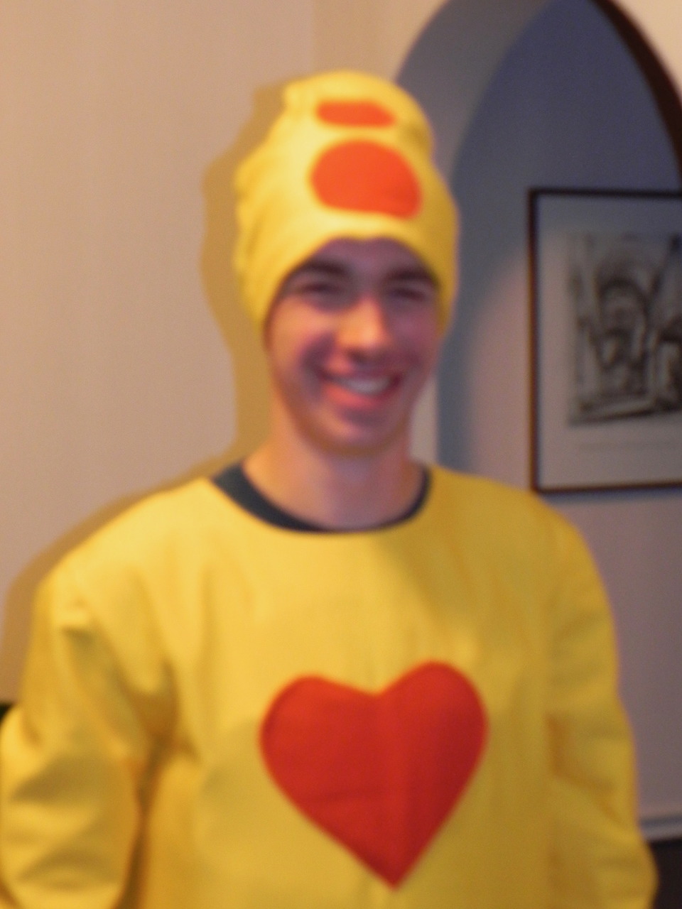 Man smiling in a yellow costume with red heart on chest and yellow hat with red dot
