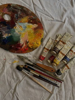 Artist palette with mixed paint, tubes of oil paint, brushes, and palette knife on white cloth