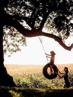 Two children in silhouette playing on tire swing under tree at sunset
