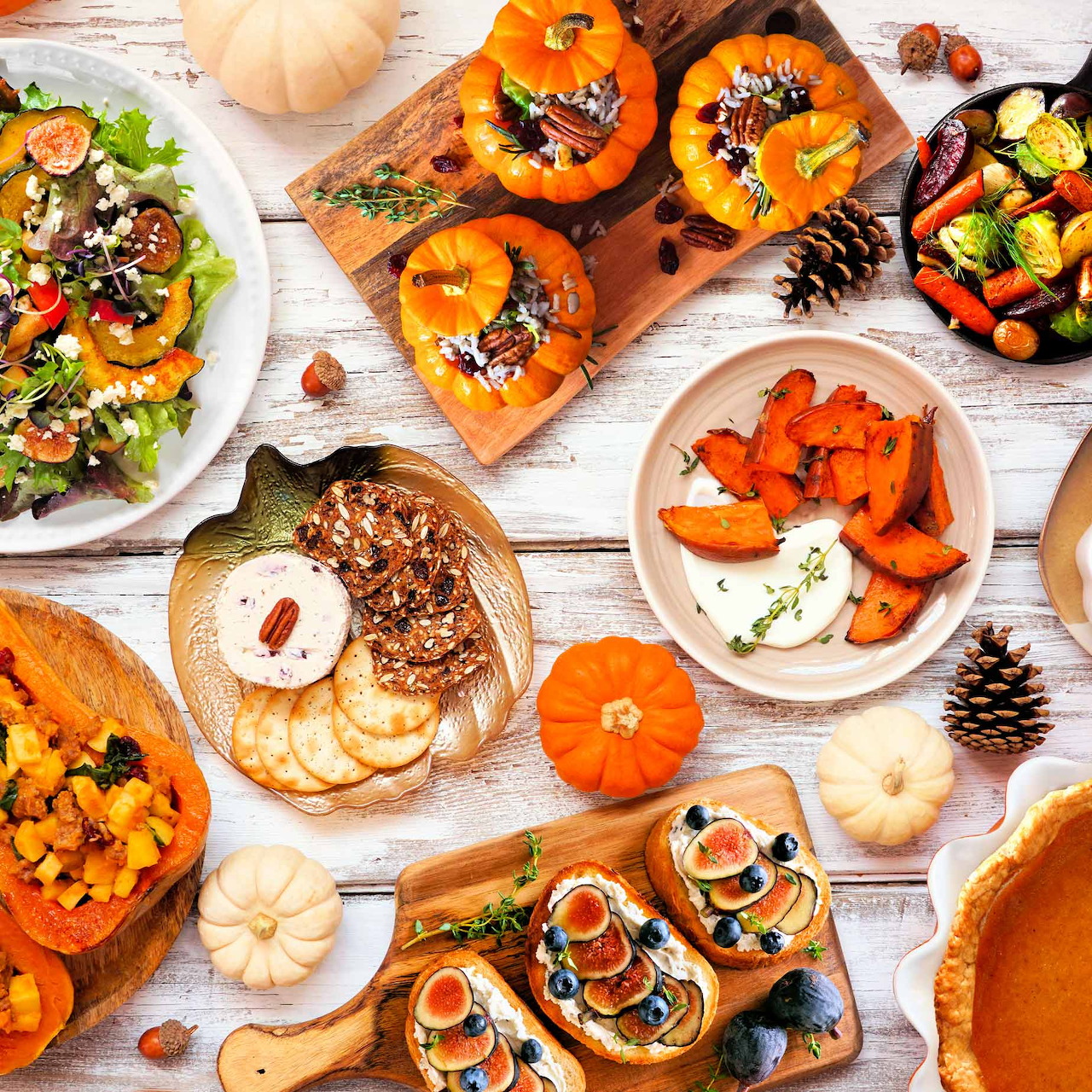 Overhead view of autumn food spread with pumpkins, roasted vegetables, salads, and desserts on white wooden table