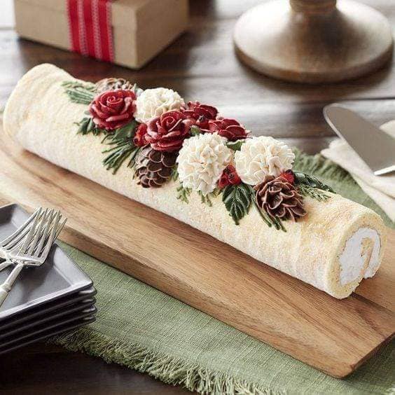 White frosted yule log cake decorated with red roses and pine cones on wooden board