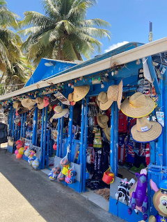 Blue-painted outdoor market stalls selling straw hats and souvenirs with palm trees and blue sky
