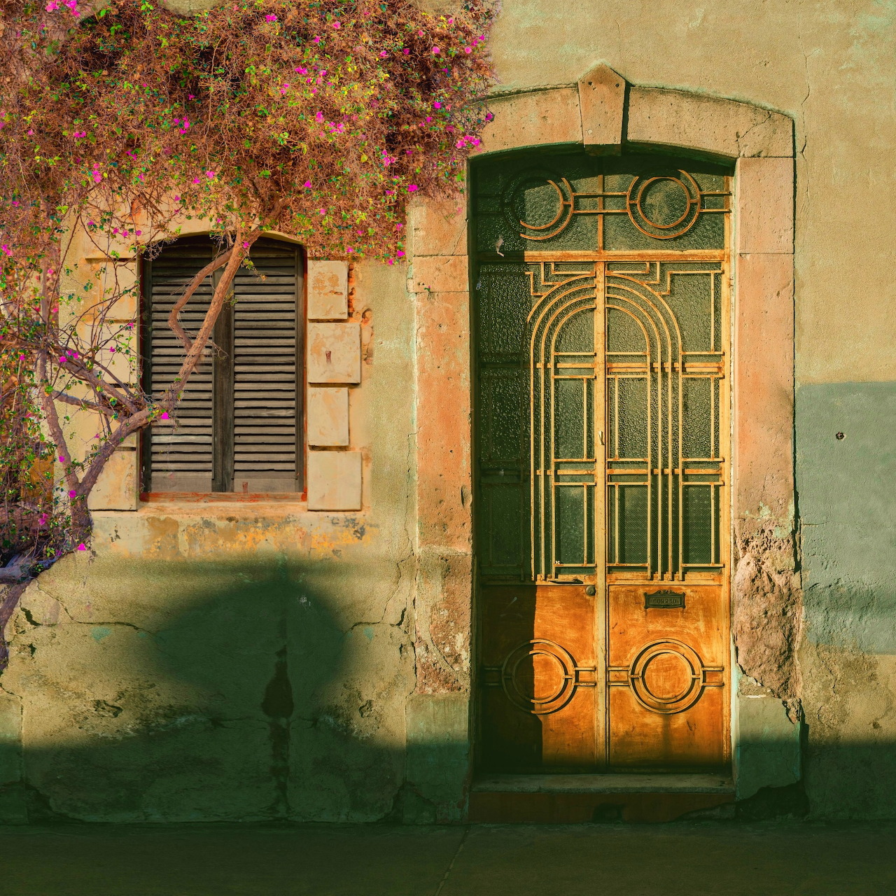 Rustic Mediterranean building with ornate door, shuttered window, and pink flowering vine on weathered wall