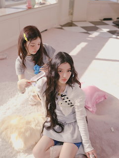 Two girls in a pastel pink room, one brushing the other's long dark hair, with a white dog resting beside them