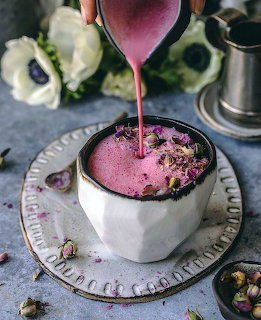 Pink latte being poured into a white cup topped with dried rose petals on a gray surface with white flowers