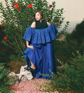 Woman in flowing blue gown posing in garden with roses and green foliage