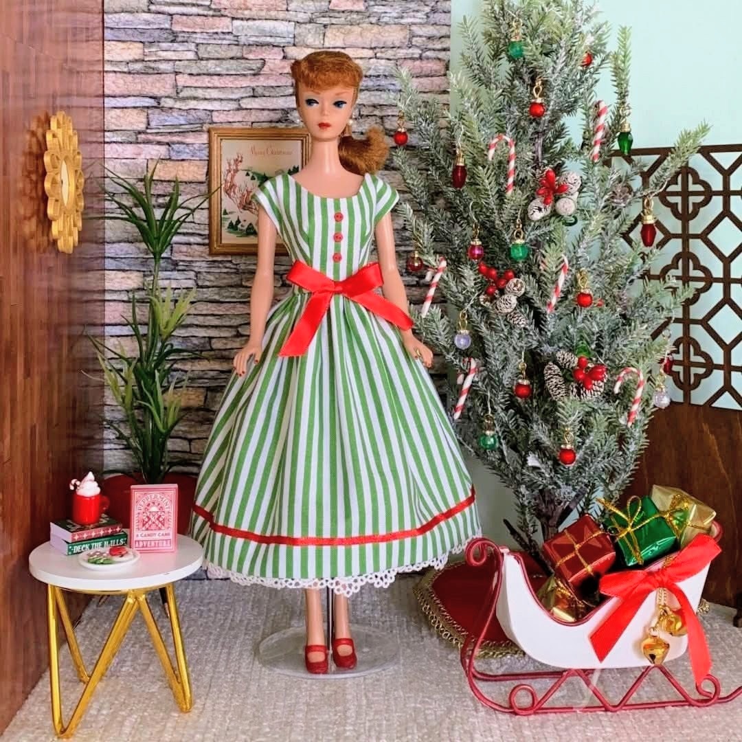 Vintage Barbie doll in green striped dress with red bow beside decorated Christmas tree and miniature furniture