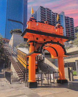 Orange funicular railway car on elevated orange arch structure with city buildings and colorful sunset sky behind