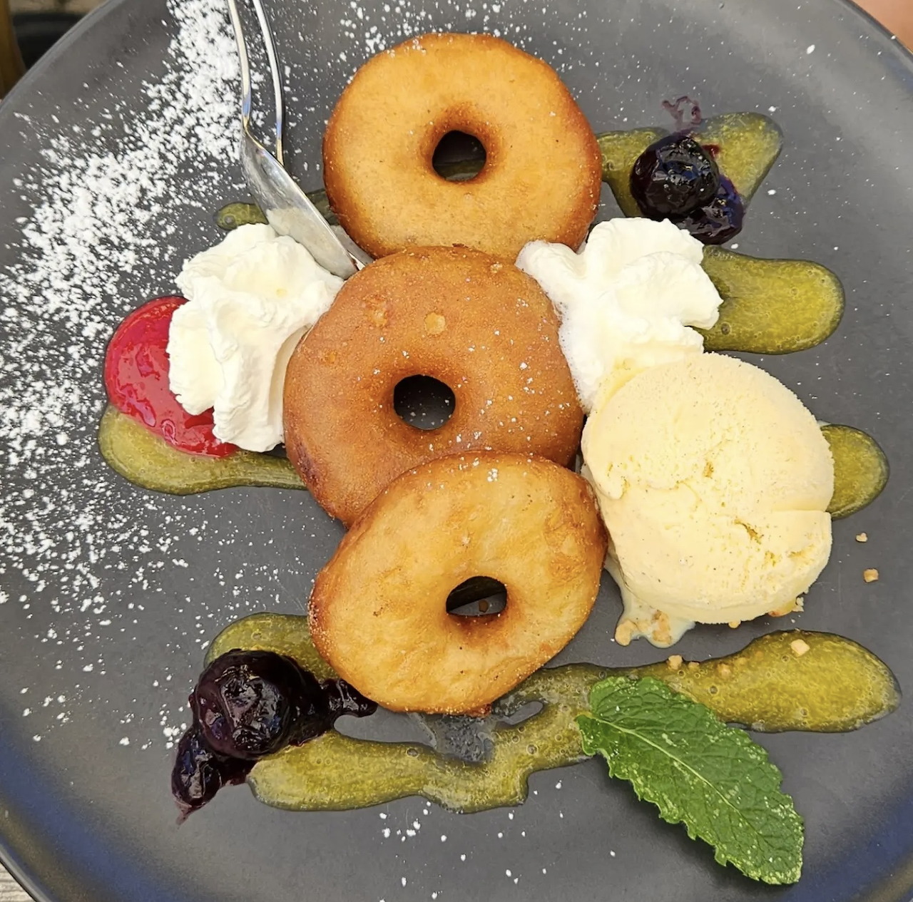 Plated dessert with donuts, ice cream, whipped cream, berries and mint garnish on dark plate with powdered sugar