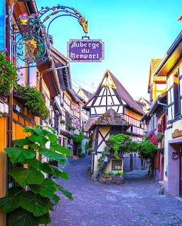 Narrow cobblestone street with half-timbered houses, flowers, and a hanging sign in a European village