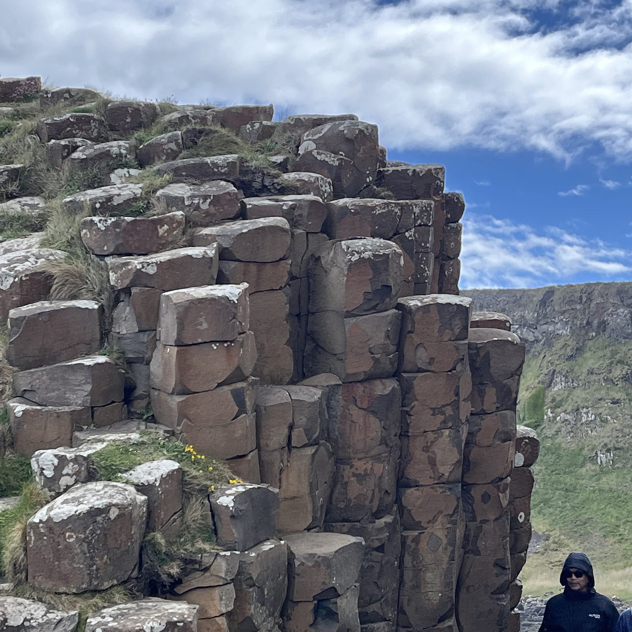 Person standing next to tall basalt rock columns under a blue sky with clouds