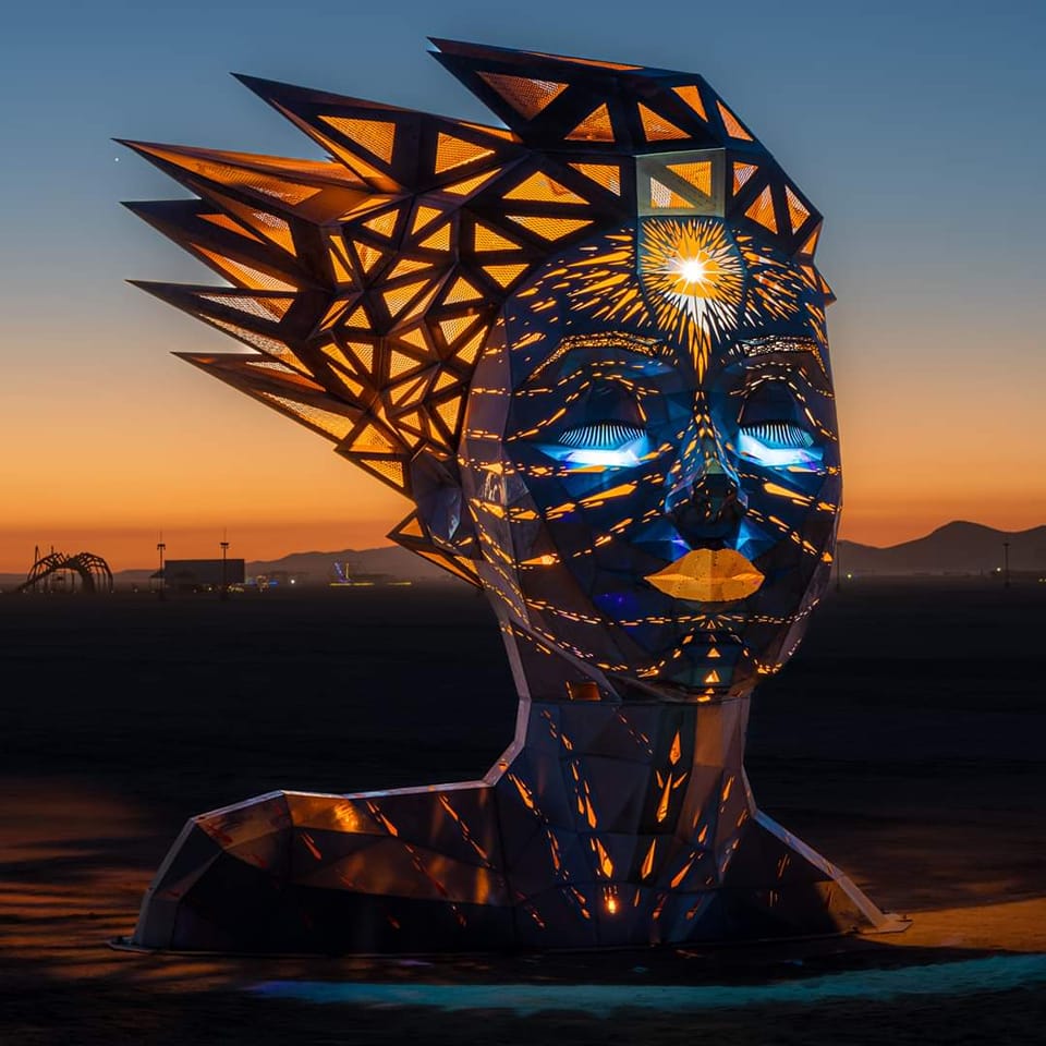 Illuminated geometric metal head sculpture at sunset with glowing interior light and desert background