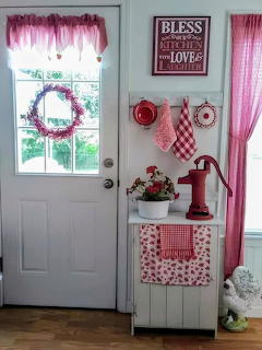 Vintage red and white kitchen corner with door, pink wreath, gingham curtains, and decorative hand pump