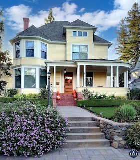 Yellow Victorian house with a wraparound porch, brick steps, hedges, and purple flowers in front