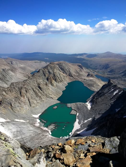 View from a rocky mountain summit over a turquoise alpine lake and rugged peaks under blue sky