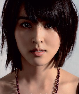Close-up portrait of a woman with short black hair and a dark beaded necklace on gray background