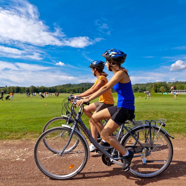 Two people riding a tandem bicycle on a dirt path through a grassy field on a sunny day