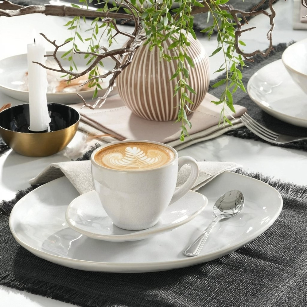 White cup of latte with leaf art on plates and a spoon on a gray table setting