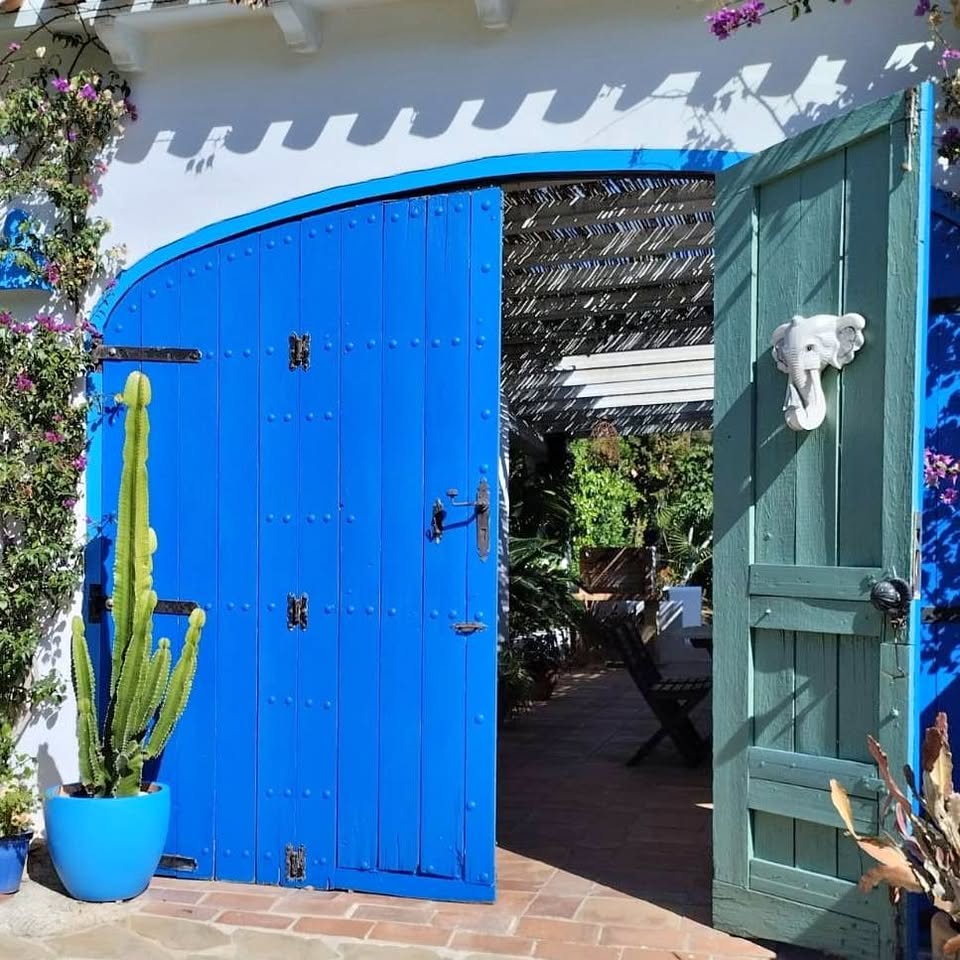 Blue arched gateway with iron grille and green door opening to sunny villa courtyard with potted cactus