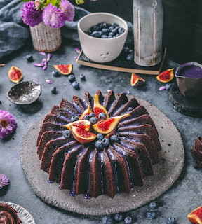 Chocolate bundt cake topped with fig slices and blueberries on slate serving board with purple flowers