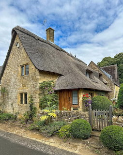 Stone thatched cottage with a garden, wooden gate, and cloudy blue sky