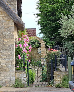 Stone cottage with black iron gate, pink flowers and evergreen trees in background