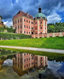 Pink castle with green copper dome reflected in a pond with manicured gardens and dramatic clouds