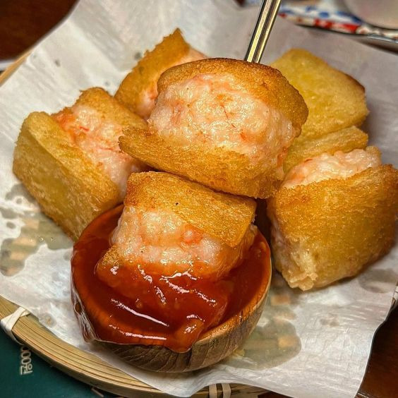 Golden fried shrimp toast pieces on white plate with red sauce in small bowl, chopsticks holding one piece