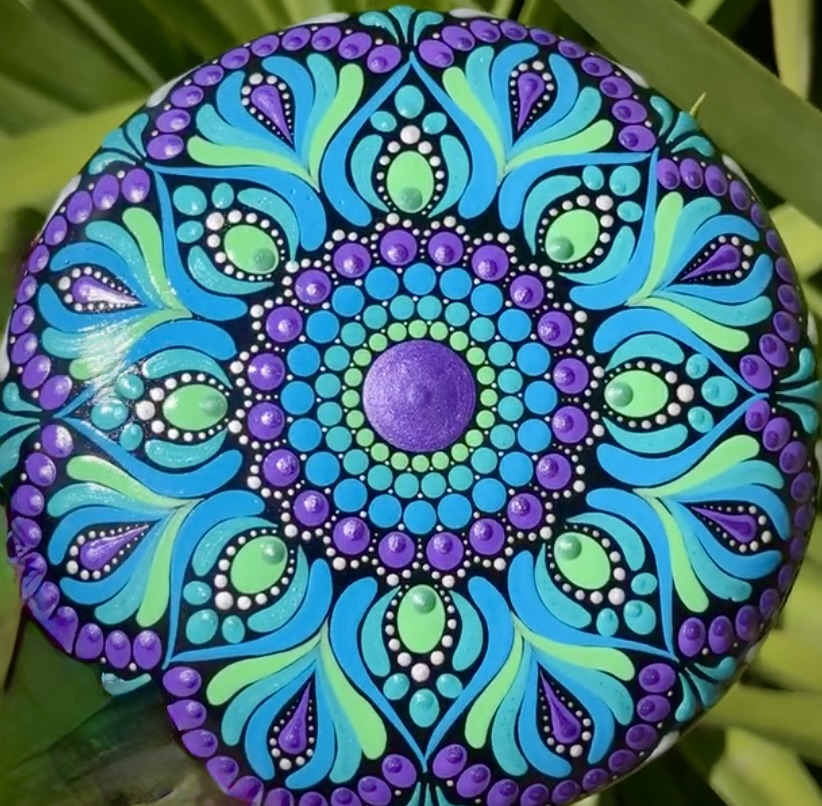 Blue and purple mandala painted on a round stone with green leaves in the background