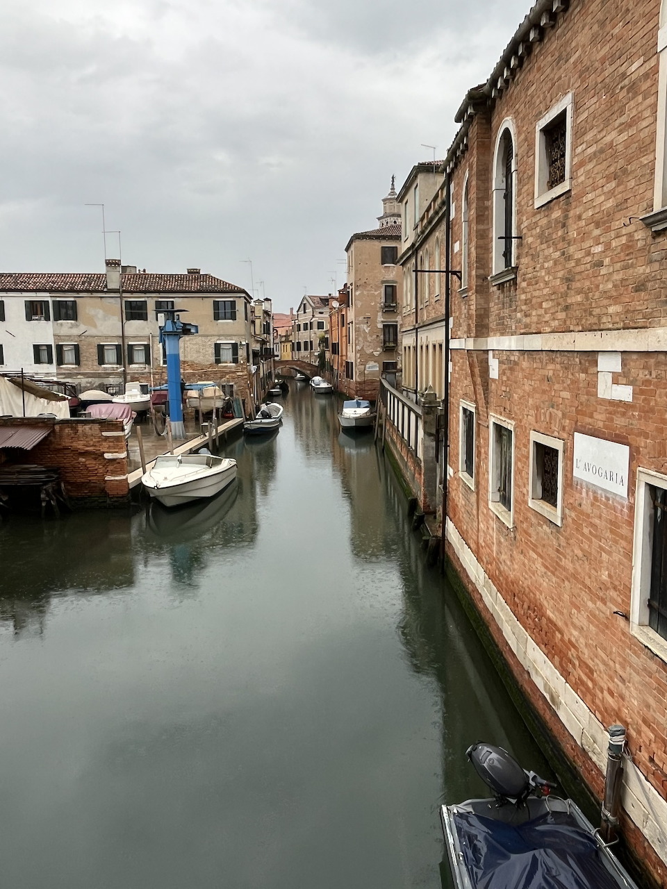 Narrow Venice canal with moored boats and brick buildings on both sides under cloudy sky
