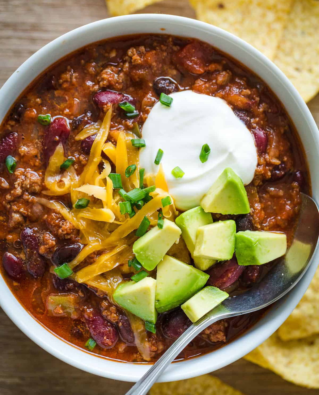 Bowl of chili with sour cream, diced avocado, shredded cheese, and chives beside tortilla chips