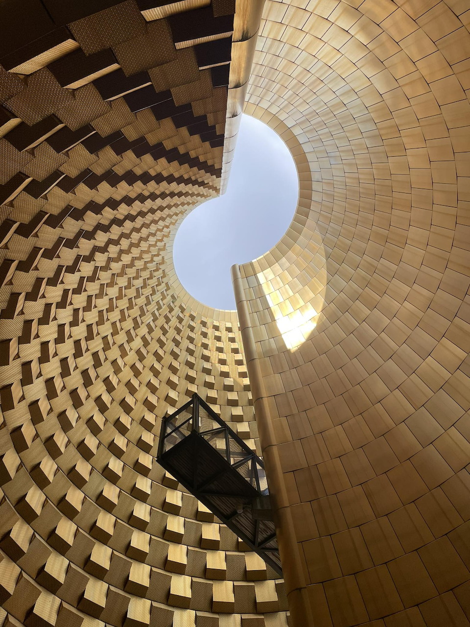 Looking up inside a circular building with patterned walls, a round skylight, and a suspended walkway