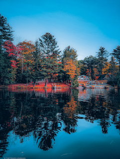 Autumn trees with orange and red leaves reflected in a still lake, small bridge visible among the trees