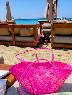 Pink woven beach bag on sand with turquoise ocean and beach umbrellas behind