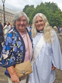 Two women standing together outdoors, one in a floral jacket and one in clergy robes, smiling at camera